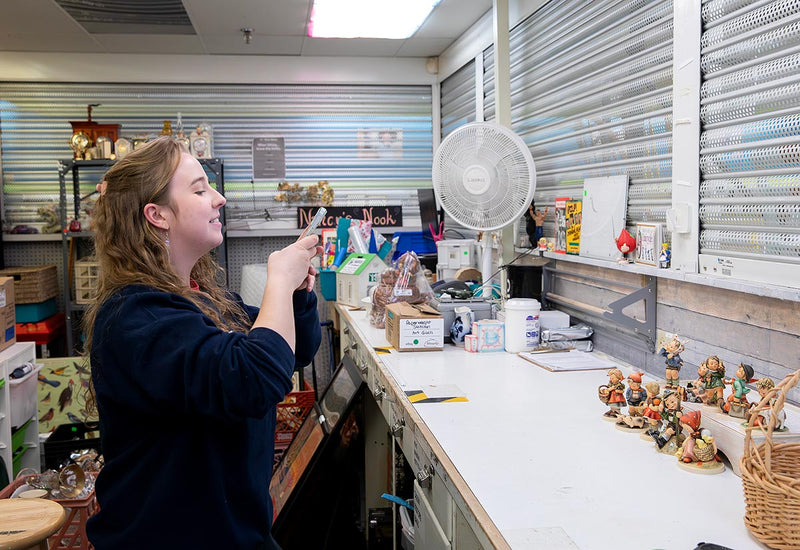 A woman photographs a shelf filled with various items, capturing the arrangement and details of the objects displayed.