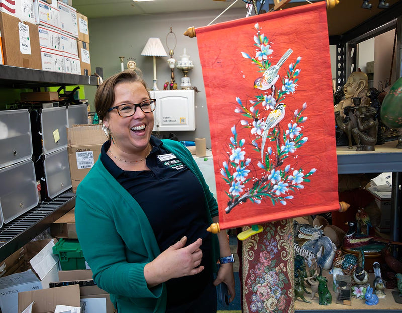 A woman in a green cardigan and black shirt laughs while holding a red scroll with a painting of birds on a flowering branch.